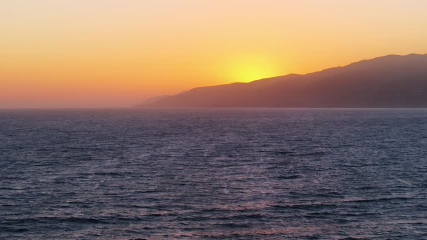 A mesmerizing aerial view of a peaceful Malibu sunset in California, USA. The hills silhouette against the golden water glow as the sun sets, creating a picturesque view over the ocean