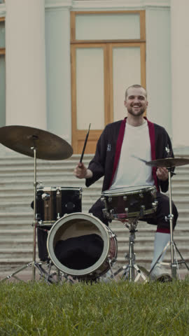 Vertical shot of cheerful male musician playing drum set with smile and adjusting cymbal during outdoor rehearsal with band