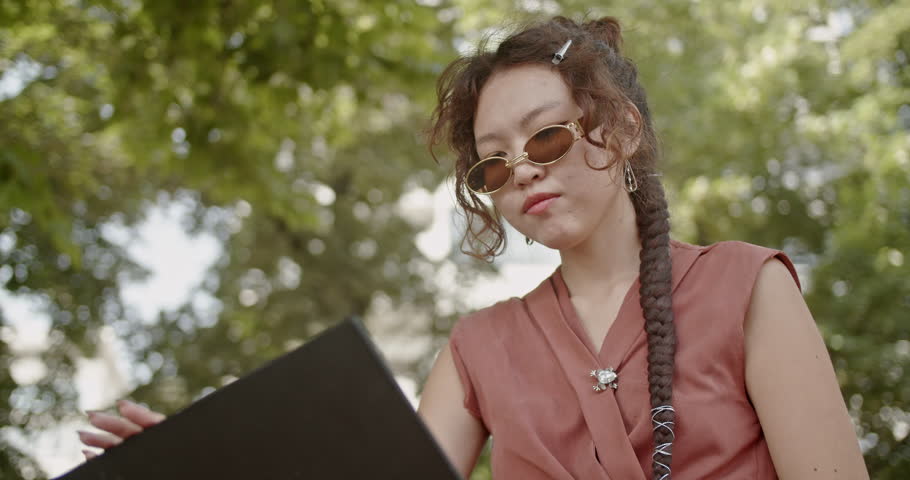Young woman wearing sunglasses and using a laptop in a park setting, working or studying outdoors.