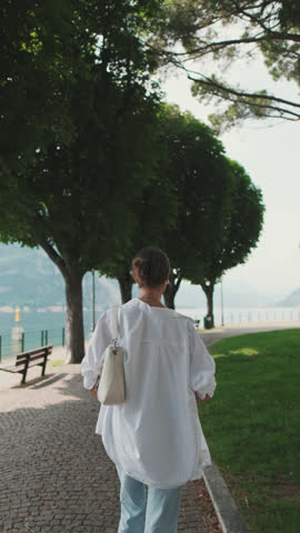 Vertical video, Young woman brown-haired dressed in white shirt, walking along the embankment of the lake of the old European city