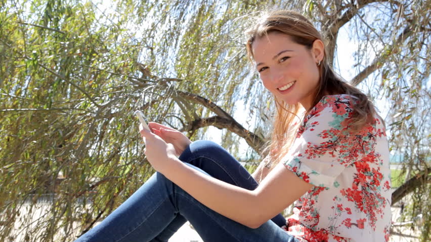 Profile portrait view of an attractive young woman using a smarphone while sitting on a stone wall in the park during a sunny breezy day, smiling.
