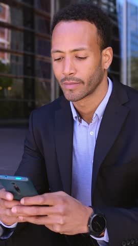 Young attractive african american business man using mobile phone sitting at staircase building entrance. Vertical HD video