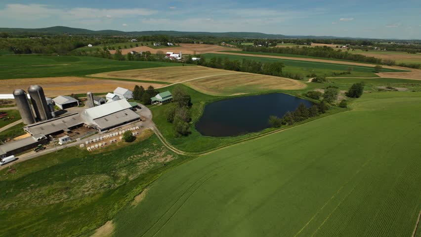 Aerial view of a serene countryside farm with silos, barns, a pond and yellow fields, surrounded by distant farms. Drone Shot in rural USA.