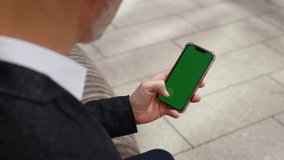 Businessman in a dark jacket and white shirt is sitting on a bench, using a smartphone with a green screen. The man is focused on his smartphone, engaging in online activities or using a mobile app. - Powered by Shutterstock - Get 15% off with code: PIKWIZARD15