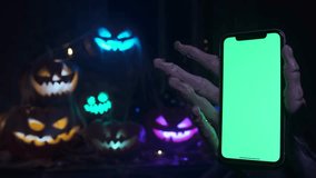 A phone with a green screen is held by a gloved skeleton hand on a Halloween background from a pile of cut-out Jack O'Lantern pumpkins at a party. Holiday offers. - Powered by Shutterstock - Get 15% off with code: PIKWIZARD15
