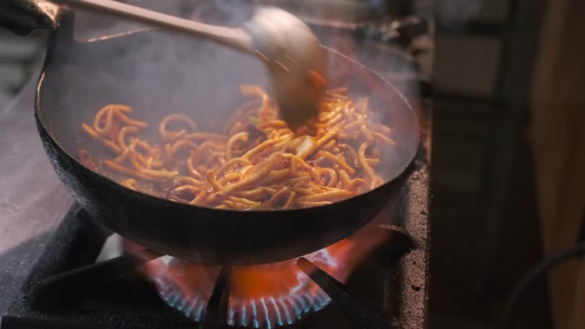 Cooking stir-fried noodles with vegetables and meat on high heat with shaking and stirring with steel ladle in commercial kitchen, close up.