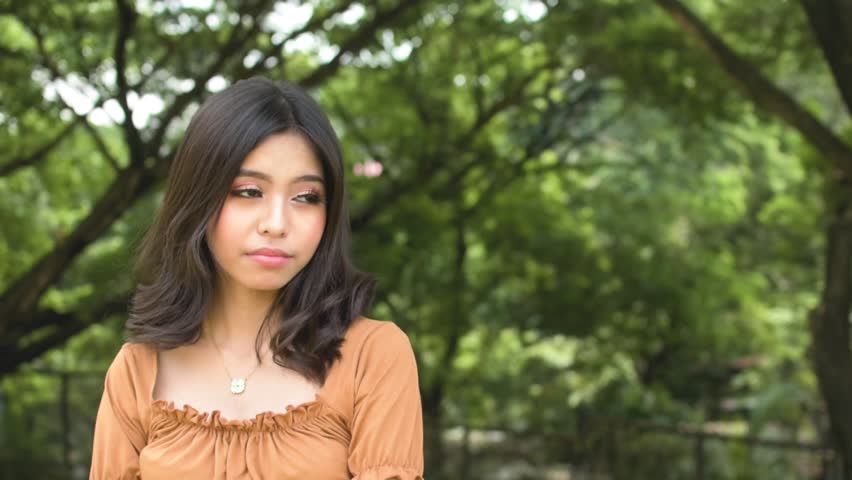 A young teenage asian woman trying to look cute for someone, smiling trying to get attention. Outdoor park scene. Copy space on the right. Slowed down 50 percent.