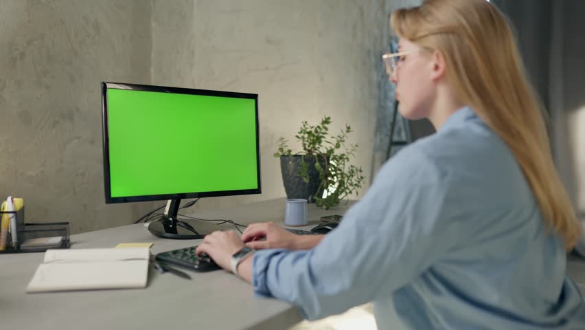 Young Woman Working In Office On Desktop Computer with Green Screen Mock Up Display. Girl Typing on Keyboard And Looks At Green Chromakey Monitor