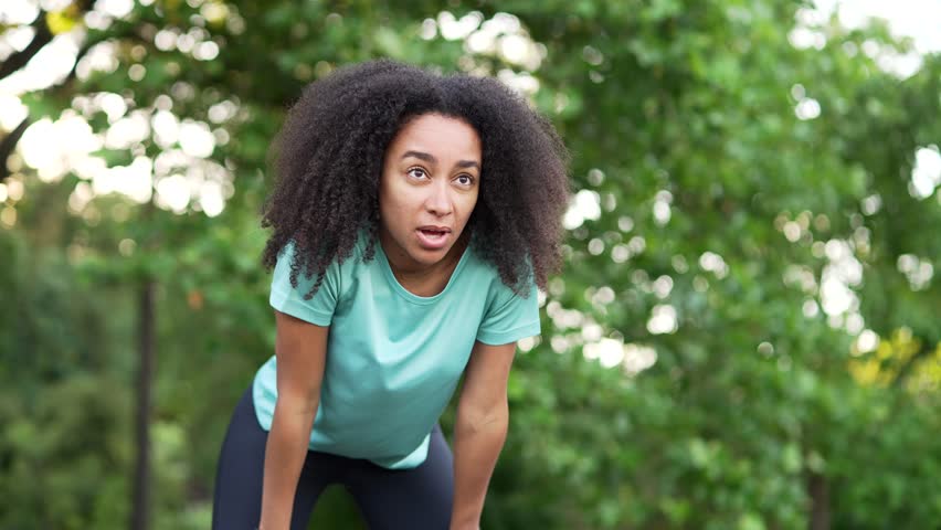 African american female runner is short of breath after a fast sprint run in urban city park. Young black woman athlete leans on her knees, wipes sweat from her forehead and breathes deeply. Endurance