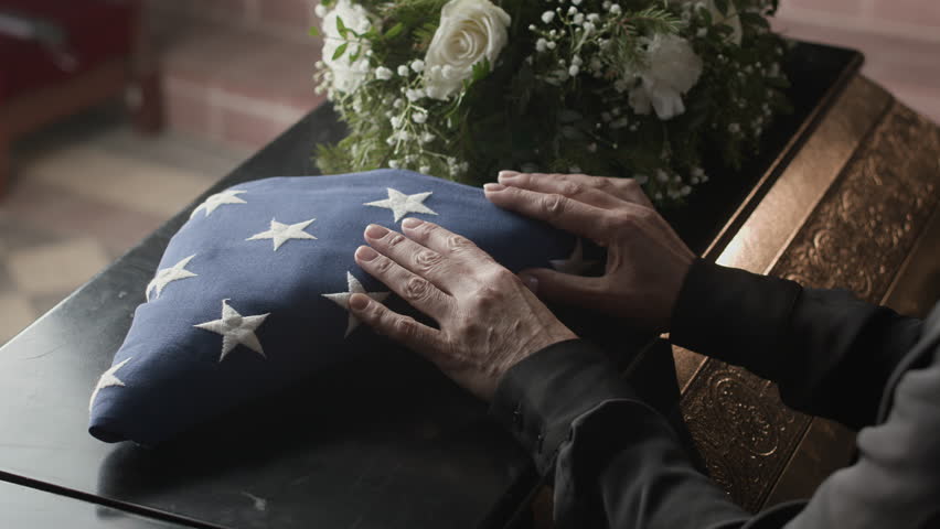 Close up shot of unrecognisable female relative putting American flag folded in triangle on coffin lid next to white flowers during memorial ceremony in church