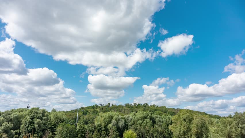 Timelapse of blue sky with white clouds floating over a hill. The hill is covered with trees and there is a cable car.