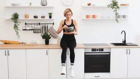 Young woman in athletic wear sitting on kitchen counter enjoying healthy salad. Bright kitchen with modern decor, houseplants, and shelves with utensils and mugs. - Powered by Shutterstock - Get 15% off with code: PIKWIZARD15