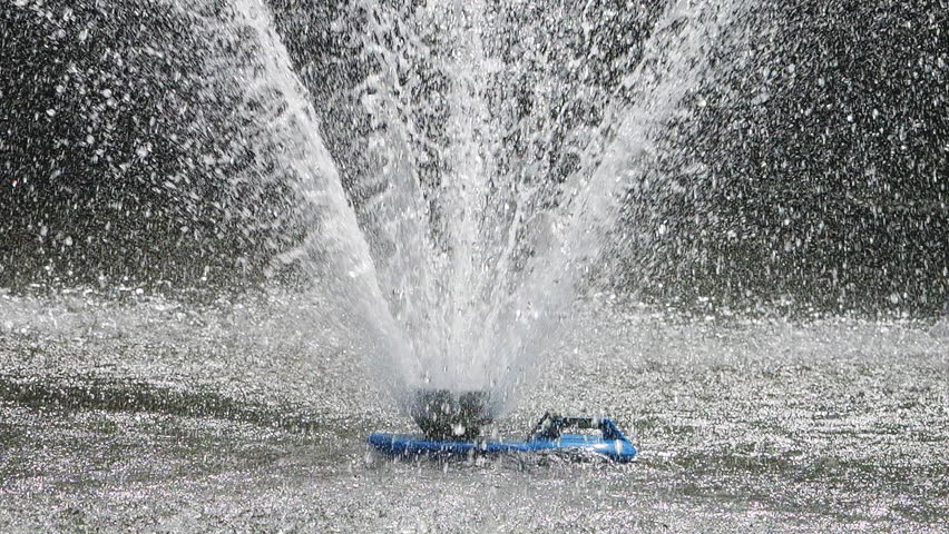 View to a pond in the park with a fountain in the middle illuminated by sunlight. Slow motion.