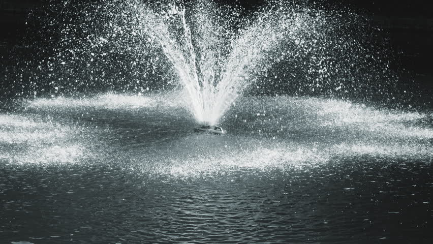 View to a pond in the park with a fountain in the middle illuminated by sunlight. Slow motion.