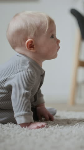 Vertical medium shot of adorable Caucasian baby girl or boy with blond hair sitting on carpet floor curiously looking around exploring surrounding space