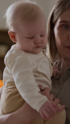 Vertical medium close-up of young Caucasian woman holding adorable baby boy or girl, lovingly kissing on cheek after daytime sleep in bedroom