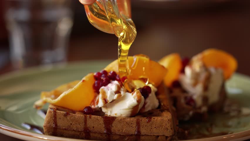 Syrup being drizzled over fresh waffles topped with cream, peaches, and berries, highlighting the rich textures. Women hands pour honey on Viennese waffles with pumpkin in a vegetarian cafe.