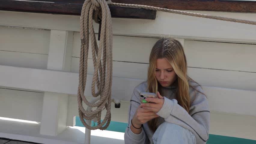 teenage girl sitting on board a ship watching a video on her phone against the backdrop of the sea