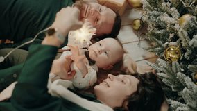 A happy family with a baby lying on the floor near a Christmas tree, enjoying the holiday together. The photo captures warmth, love, and the joy of Christmas. Happy Family Celebrating New Year - Powered by Shutterstock - Get 15% off with code: PIKWIZARD15