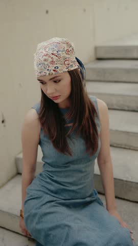 Portrait of a young woman with a headscarf, dressed in a blue dress, outdoors and seated. Captures a calm and introspective mood. Young Woman in Blue Dress with Headscarf, Outdoor Portrait