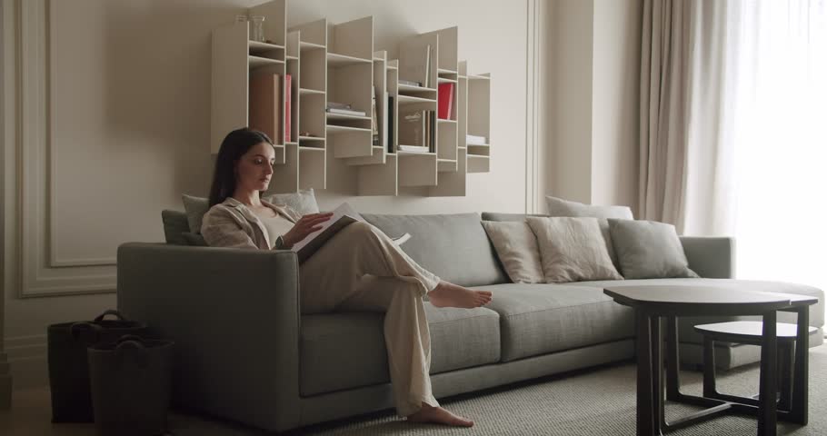 A woman sits comfortably on a modern, minimalist couch reading a book. The living room features neutral tones and stylish decor, promoting a calm and cozy atmosphere. Woman Reading Book
