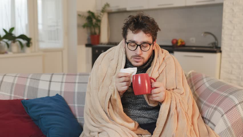 a male enduring the flu, seeking solace at home. With a cup in hand, he drinks the remedy, shivering under a duvet in the cold living room, epitomizing the harsh reality of seasonal illness