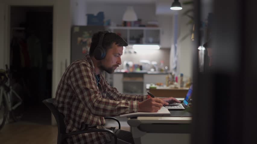 Theme of remote education. American student studying online using a laptop and wireless headphones at home. A man watches an online learning video and takes notes with a pen in a notebook. 