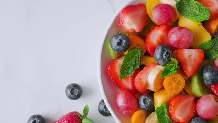 Healthy fresh fruit salad in a bowl on pink background. Top view. Summer healthy food for breakfast. Mixed tropical fruits berries for vegan diet lunch. Close up