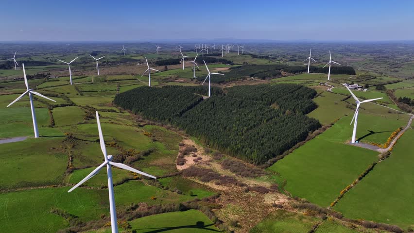 Wind Turbines, Cornasaus, County Cavan, Ireland, April 2023. Drone pulls backwards with a wide view of windmills spinning over green farmland during a clear warm morning.