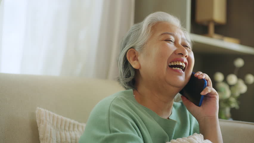 Happy Asian senior mature woman relaxing on sofa in living room speaking on mobile phone with family. Elderly grandmother enjoy smart indoor lifestyle with wireless technology and digital device.
