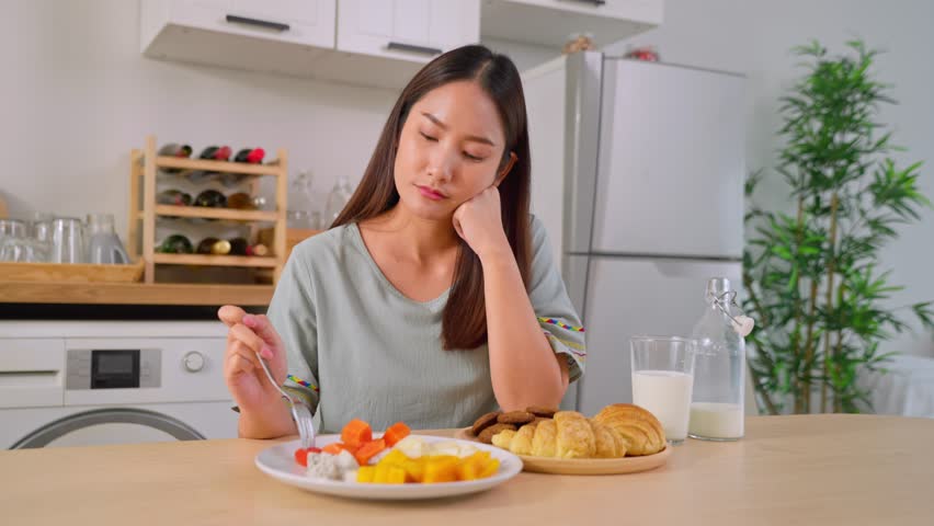 Asian unhappy woman on dieting, don't want to eat food in kitchen at home. Attractive young girl sitting on table feeling tired and bored of eating nutrition of clean healthy foods in morning in house