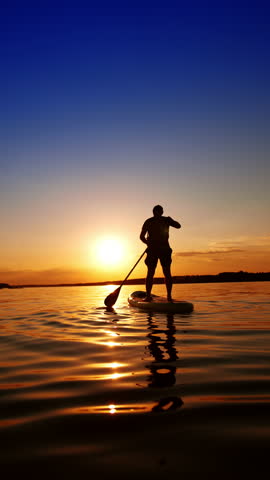 Practicing paddle boarding on the calm river in summer. Male silhouette stands on the sup board rowing with an oar. Low angle view. Vertical video.