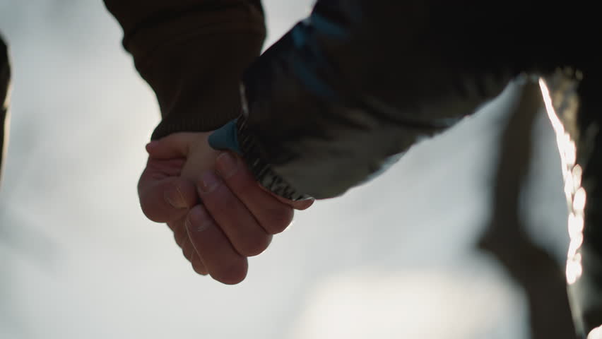 Adult and a child holding hands, illuminated by the soft glow of sunlight as he gently squeeze the child hand, with blurred view of a tree