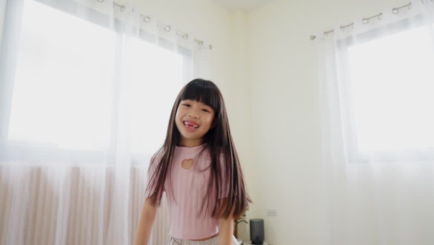 A young girl is smiling and dancing in a room with white walls and curtains. The room is well lit and the girl is wearing a pink shirt