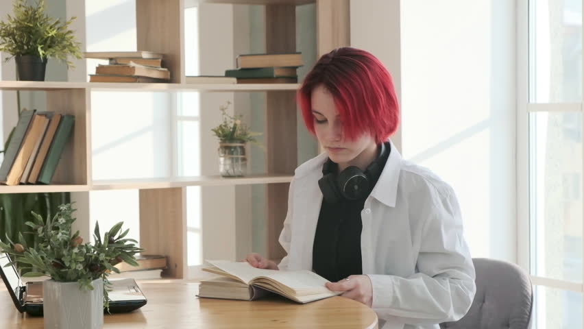 Girl with red hair is sitting at desk in a room. student is reading book and taking notes on laptop. person is reading book. home lessons