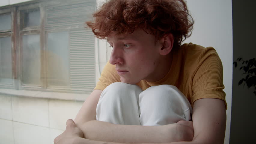 Chest up shot of young Caucasian guy with red curly hair in casual clothes sitting on window sill and looking out of window, looking sad and distressed, feeling guilty and regretful at home