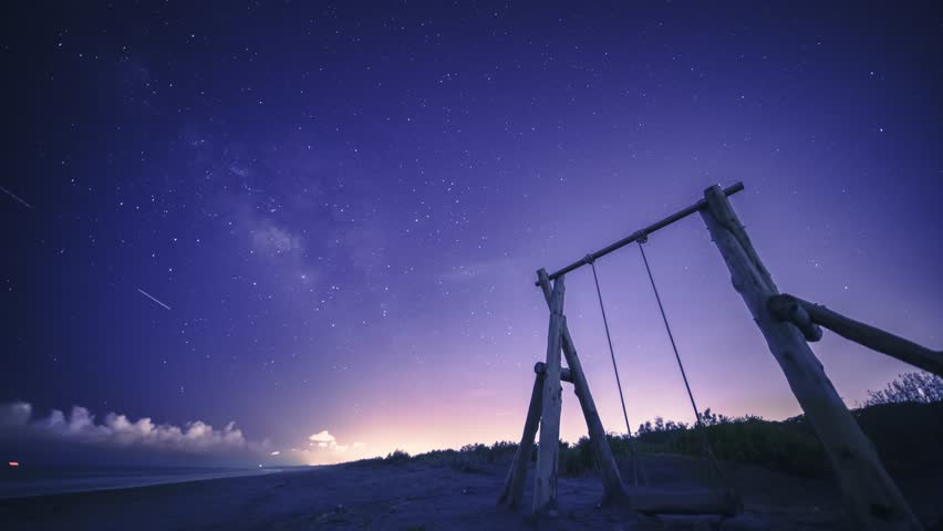 The Milky Way stretches across the night sky. A solitary wooden swing set stands on a sandy beach, inviting viewers to imagine swinging under the vast, starry sky. Zhuangwei, Yilan County, Taiwan.