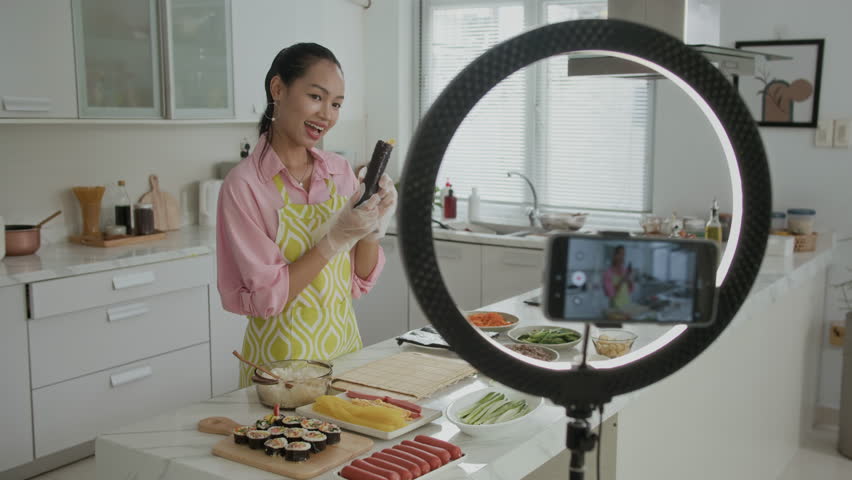 Medium long shot of gen Z Asian girl vlogging using smartphone and ring light while preparing gimbap at home kitchen