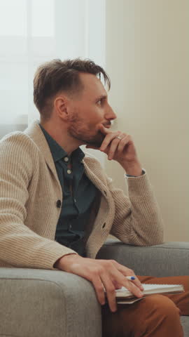 Man sits on sofa, attentively taking notes during counseling session. His focused expression suggests deep engagement with process. Concept of active participation and thoughtful reflection in therapy