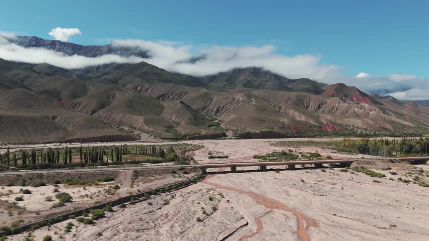 Slow pan shot on the bridge over dried river with a car driving, cloudy mountains in the background, province of jujuy in argentina