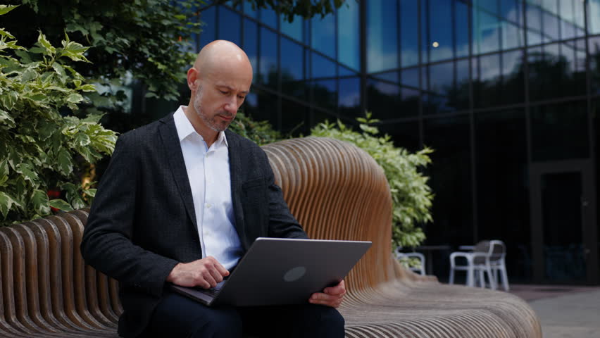 A businessman dressed in a white shirt and dark blazer is sitting on a modern bench. The man is using a laptop, having a video call with his work colleagues.