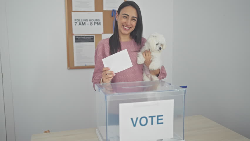 A smiling hispanic woman holding a maltese dog casts her ballot in an election at an indoor voting booth.