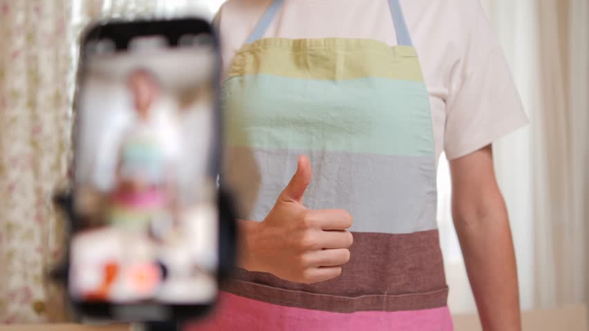 Person in kitchen apron giving thumbs up in front of smartphone camera setup