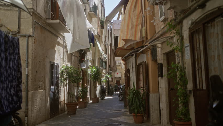 A young hispanic woman dressed in white walks down the charming old town streets of bari, italy, with potted plants, hanging laundry, and rustic buildings lining the narrow european alley.