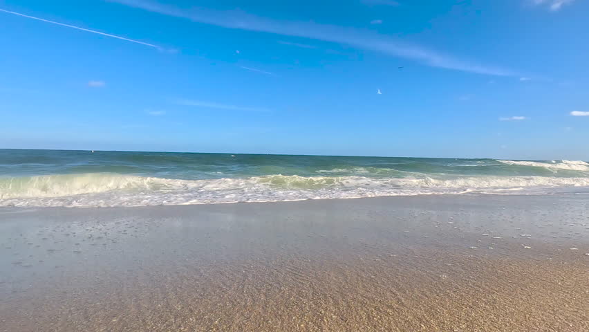 Slow motion low-angle shot tracks toward ocean surf, as foamy waves splash over top of camera lens on Ft. Lauderdale beach.