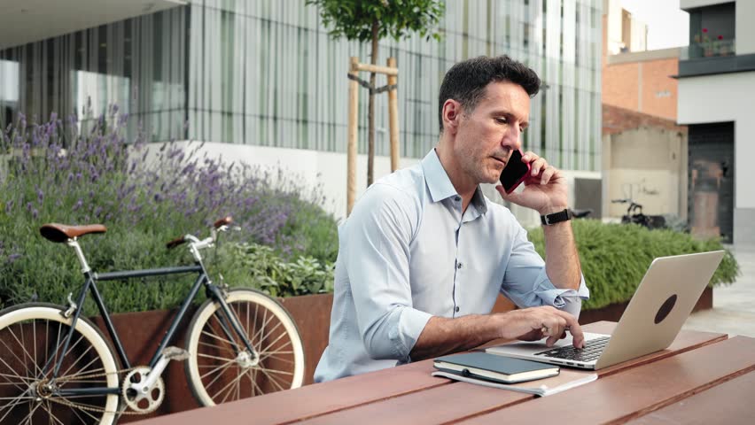 Professional man on a cell phone call using laptop outside the office, talking about business and freelance projects