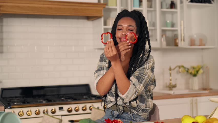 Portrait of funny black girl playing with slices of bell peppers in kitchen. Happy young vegetarian woman putting paprika as goggles, cooking at home and having fun, joking with vegetables, good mood