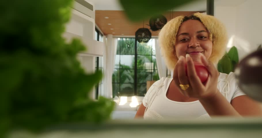 Young African American plus size woman opens refrigerator door and chooses healthy food with vegetables. View from inside the refrigerator. Diet and weight loss. Body positive.