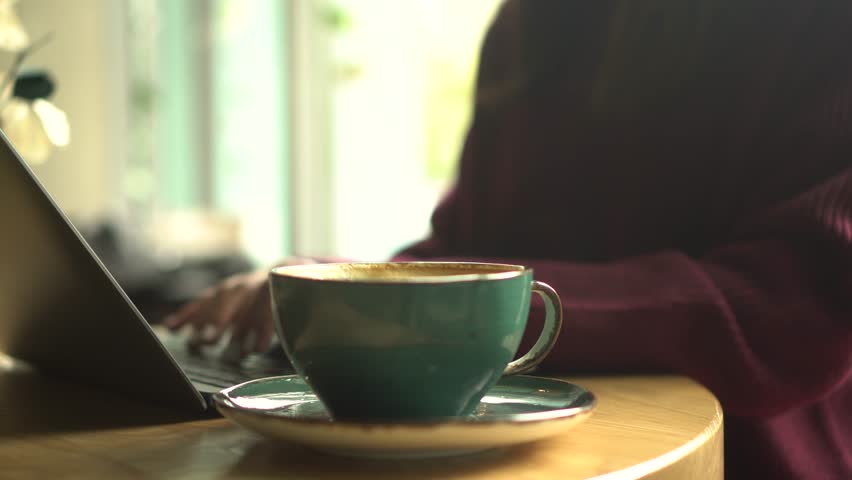 A young woman is working on a laptop and drinking coffee in a cozy cafe with a panoramic window in the background.
