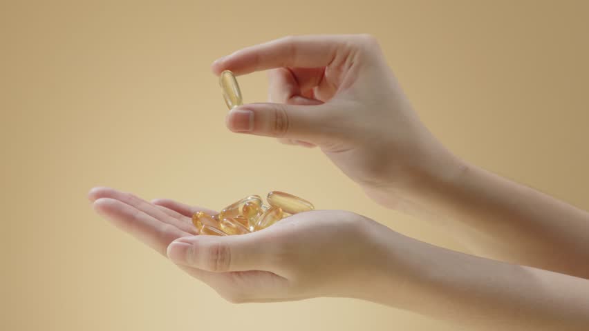 Front close up shot at two beautiful hands with yellow soft gel supplement capsules held in palm and being kept by two finger. Beige background commercial footage, empty space for text